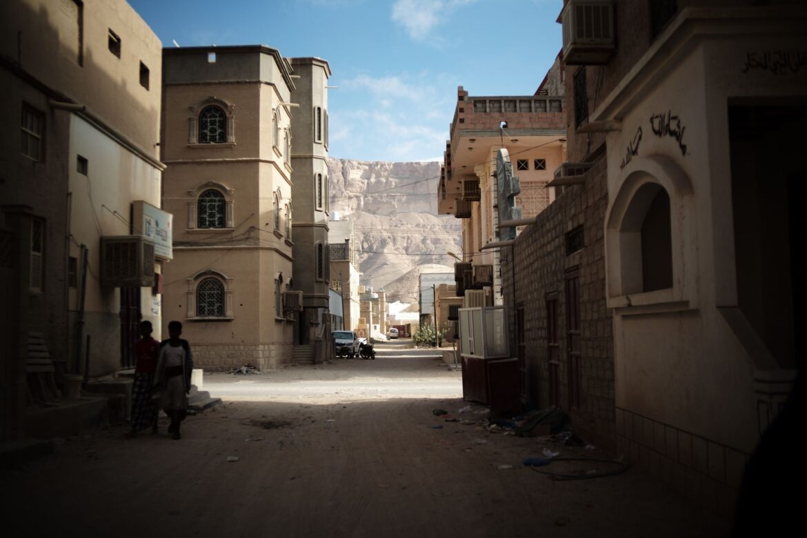 Una calle tranquila en un pueblo de Oriente Medio, enmarcada por arquitectura tradicional y con un fondo montañoso.