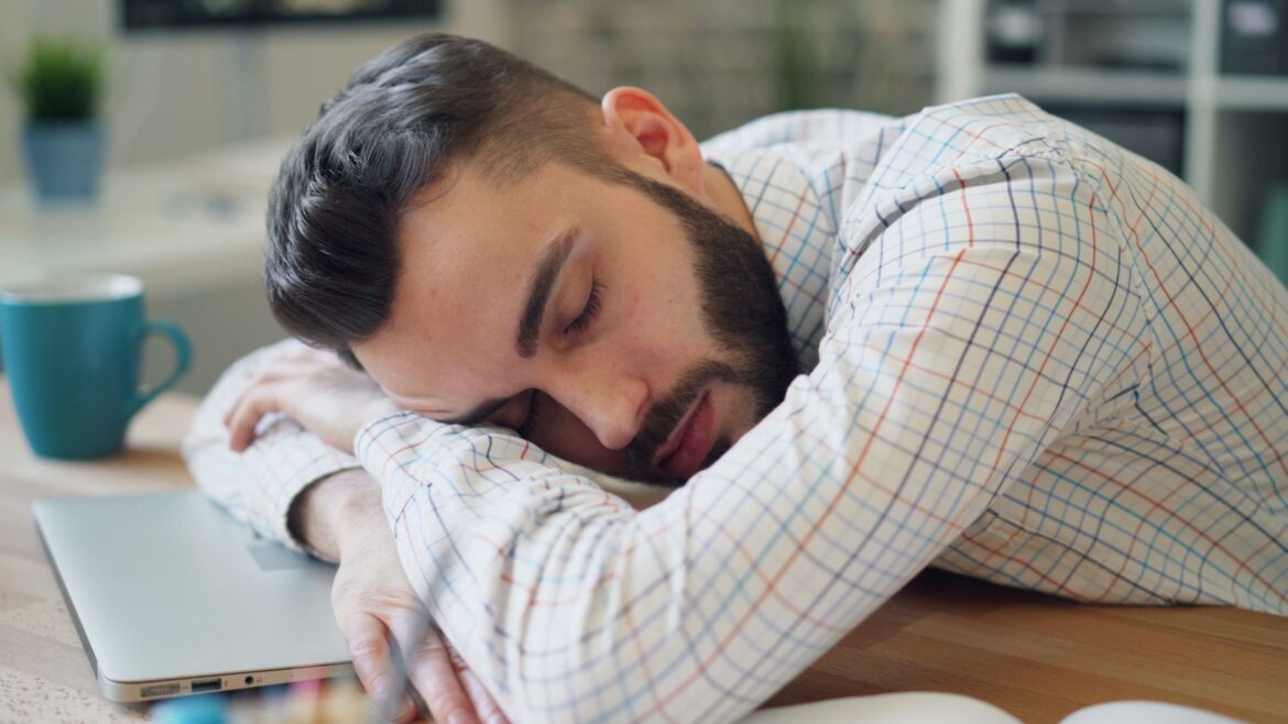 Hombre con barba descansando en un escritorio junto a un portátil, aparentando estar exhausto en un entorno de oficina.