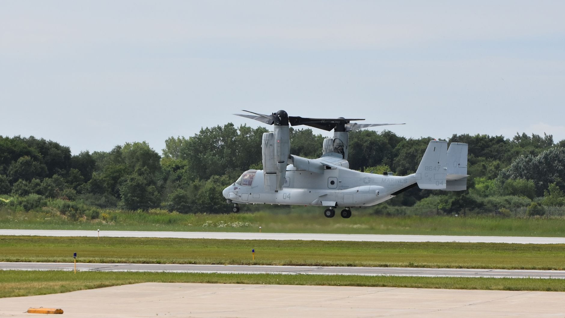 Una aeronave militar MV-22 Osprey aterrizando en la pista de un aeródromo boscoso.