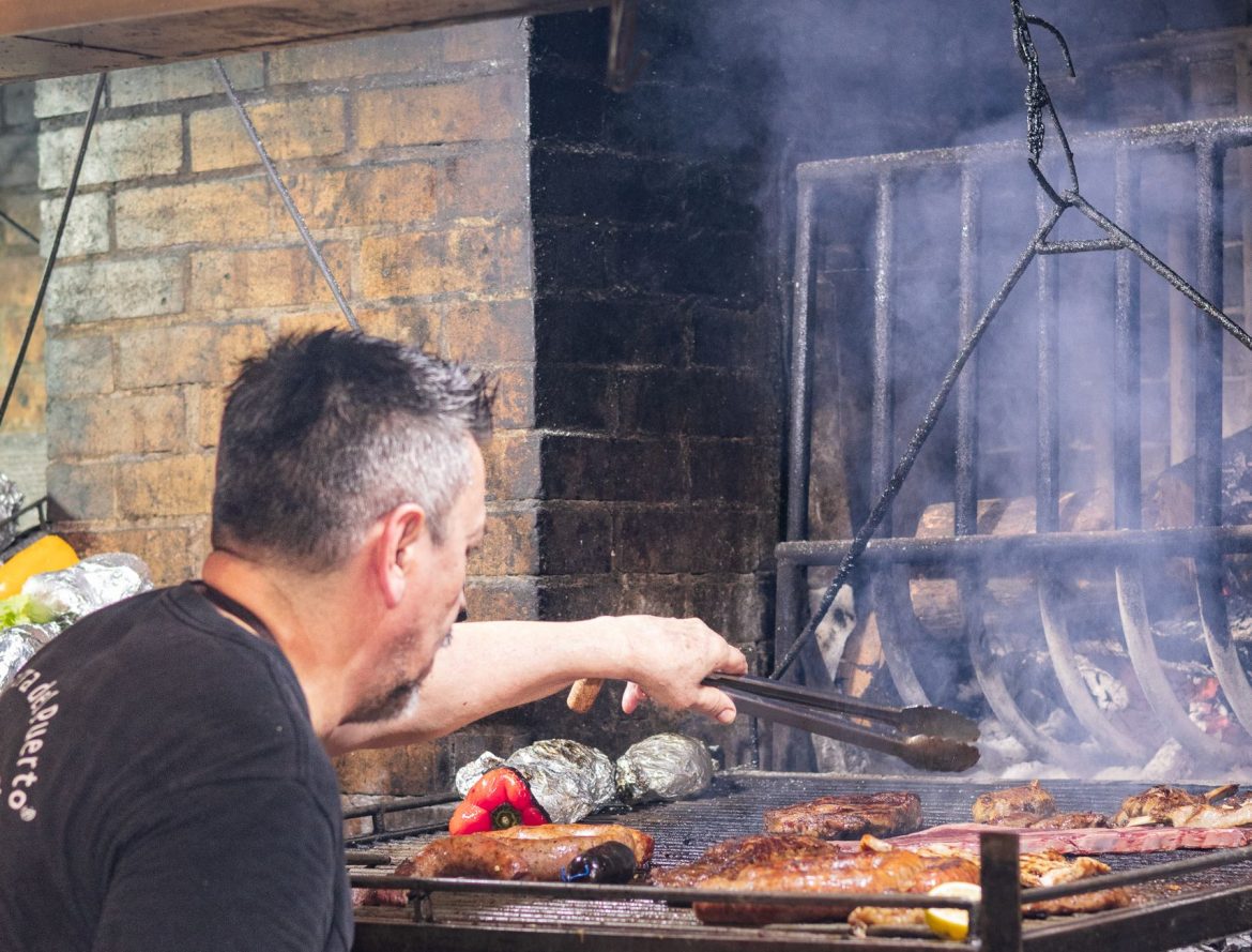 Un hombre asa a la parrilla, sobre fuego abierto, un asado tradicional uruguayo que incluye carnes y verduras.