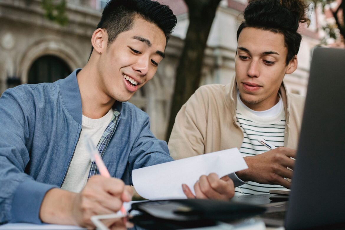 Dos estudiantes universitarios participando en una sesión de estudio al aire libre, discutiendo y escribiendo en documentos.