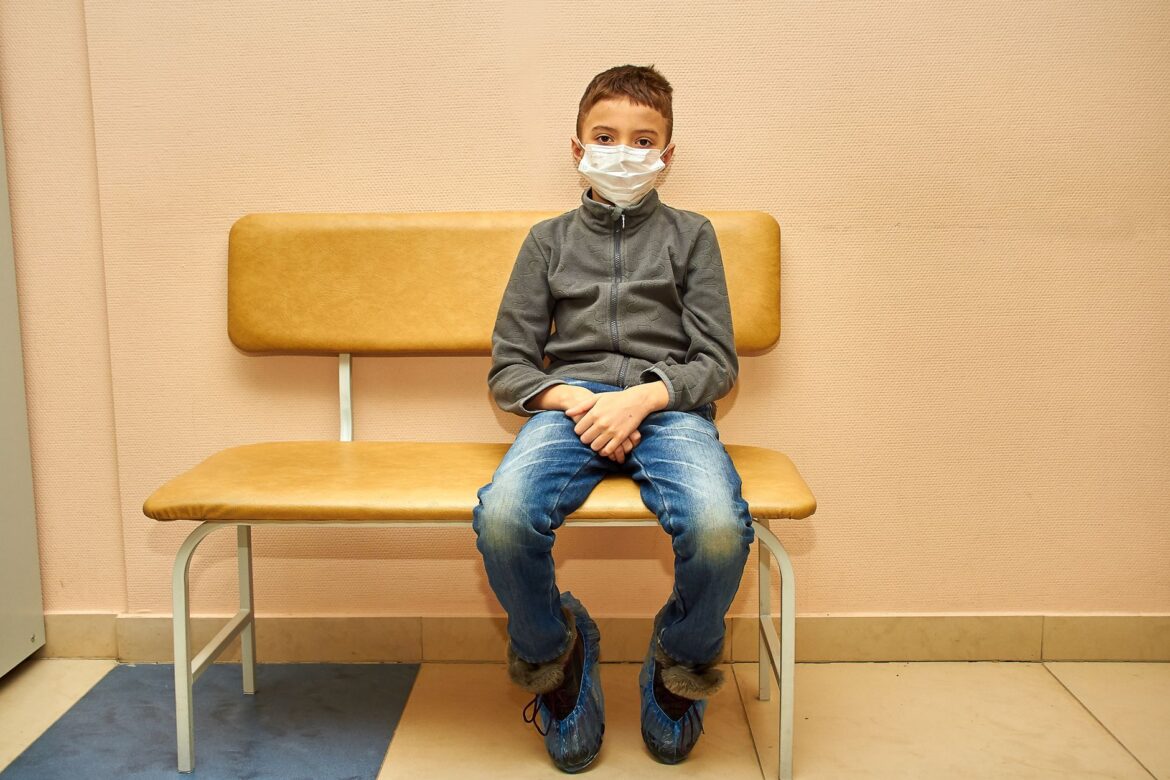Niño pequeño con mascarilla sentado en un banco, destacando la nueva normalidad en entornos de atención médica.