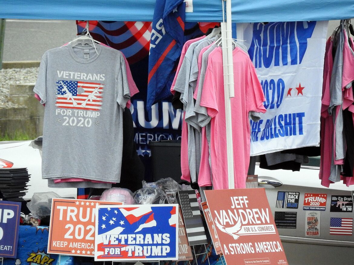 Un puesto que muestra productos de Trump 2020, incluyendo camisetas y carteles en un mercado al aire libre.